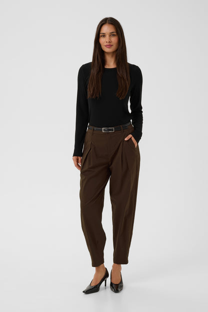 A woman modeling a black long-sleeve top and dark brown pleated trousers with a black belt, paired with black heels, against a light gray studio background.