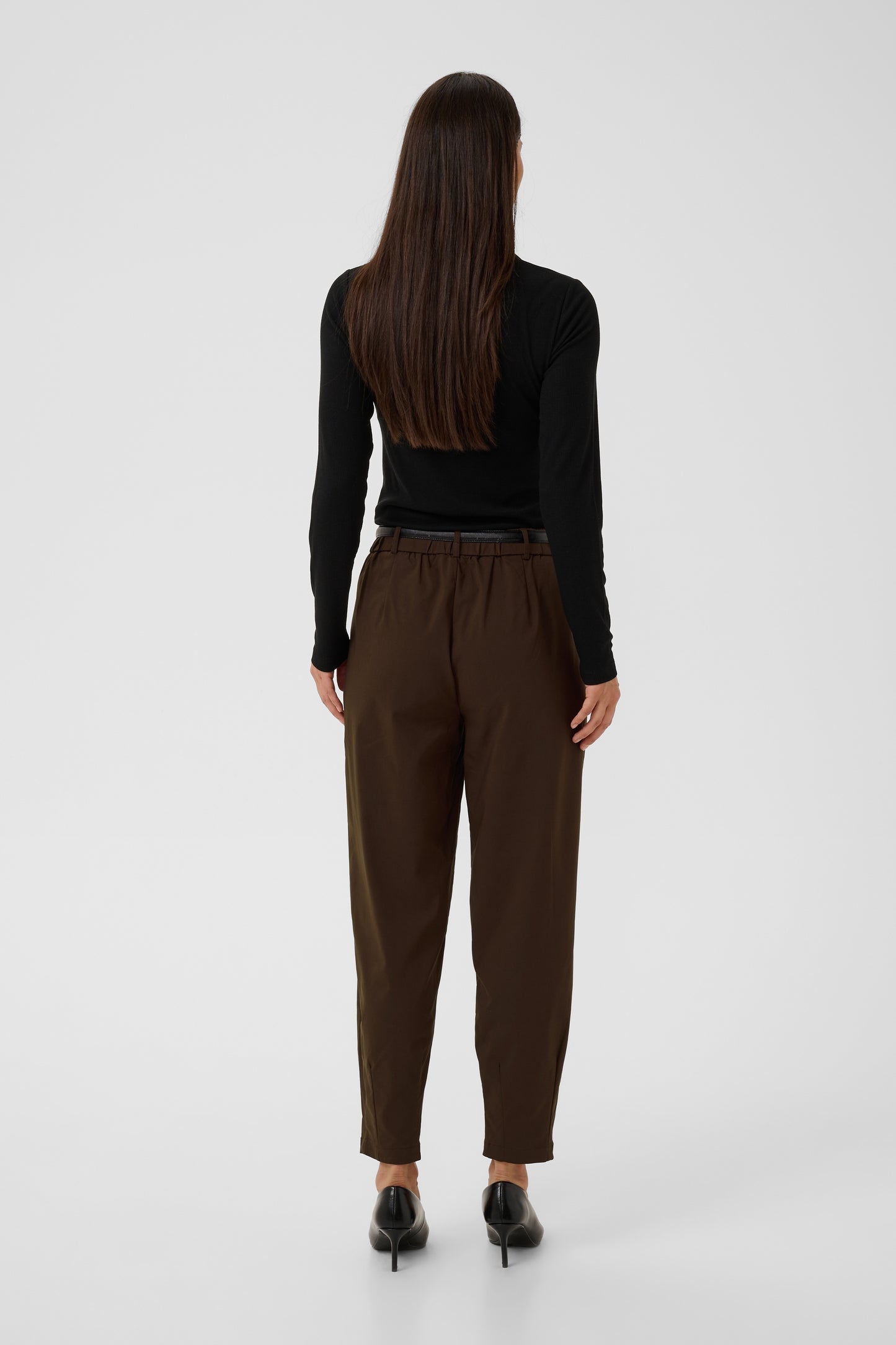 A woman viewed from behind wearing dark brown trousers, a black long-sleeve top, and black pointed shoes against a light gray studio background.