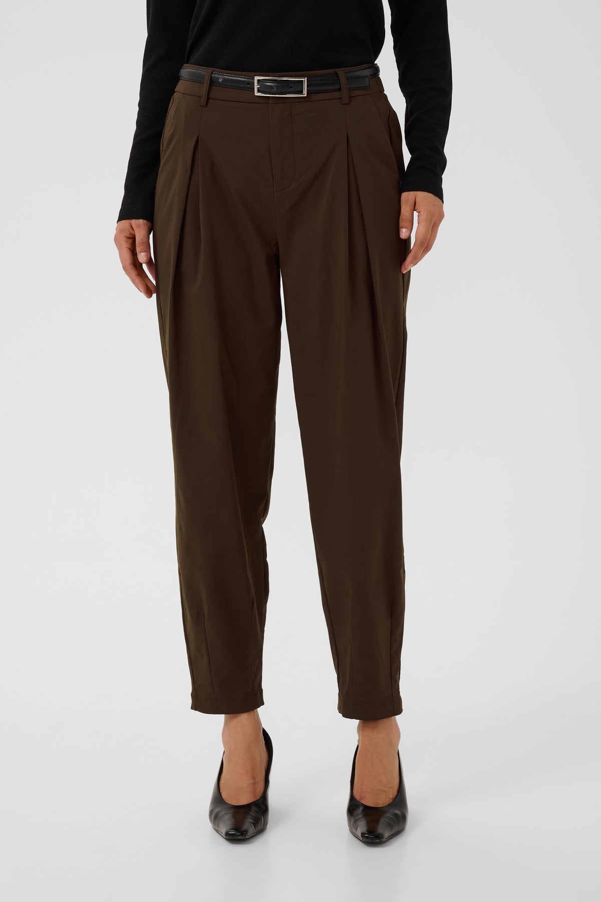 A person wearing dark brown pleated cropped trousers with a black belt, black long-sleeve top, and black pointed shoes, photographed against a light gray studio background.