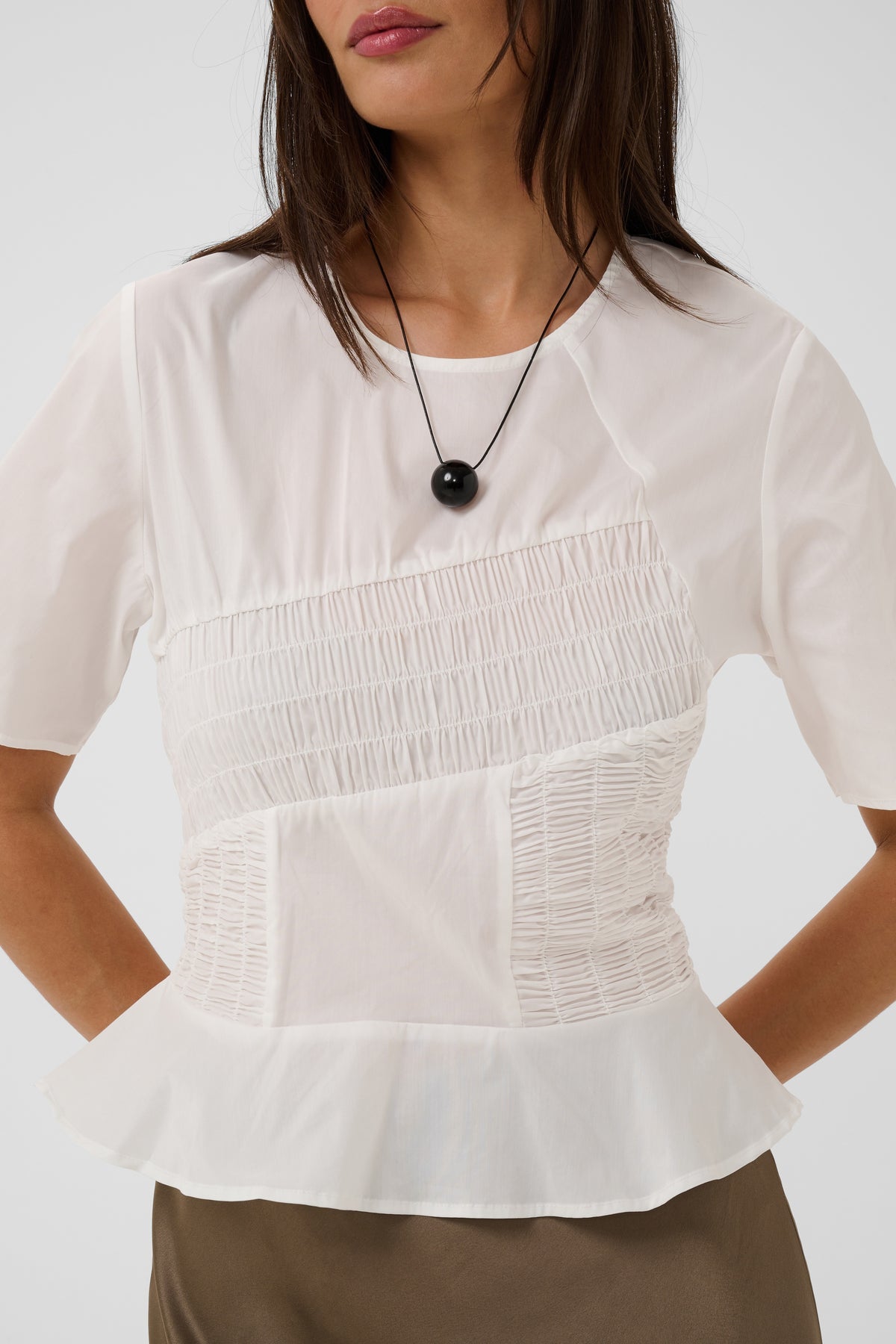 A woman wearing a white short-sleeve blouse with smocked detailing across the chest and a ruffled peplum hem, paired with brown bottoms and a black pendant necklace.