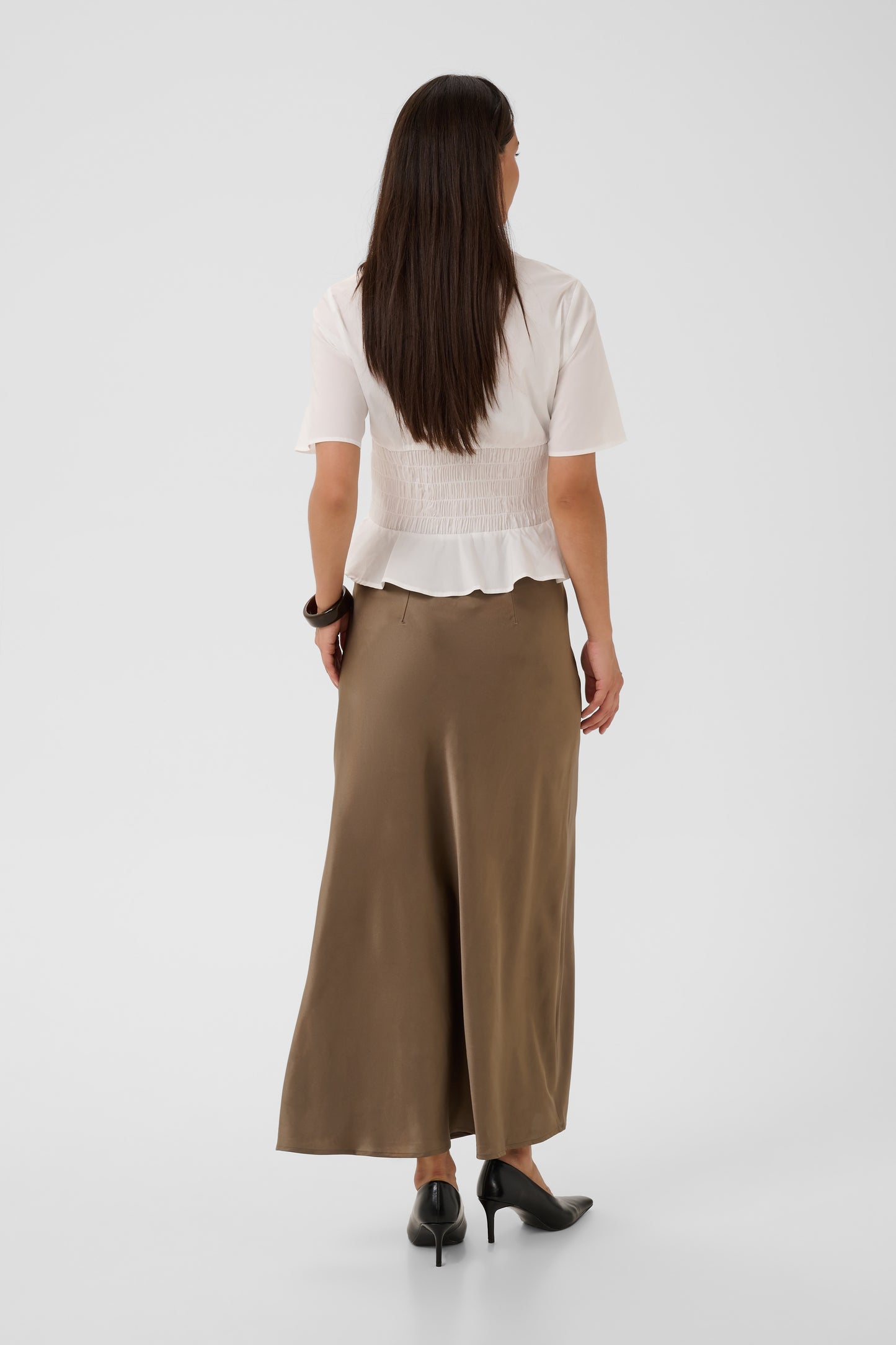 A woman shown from the back wearing a white smocked short-sleeve blouse and a brown wide-leg midi skirt, paired with black heels, against a light gray studio background.