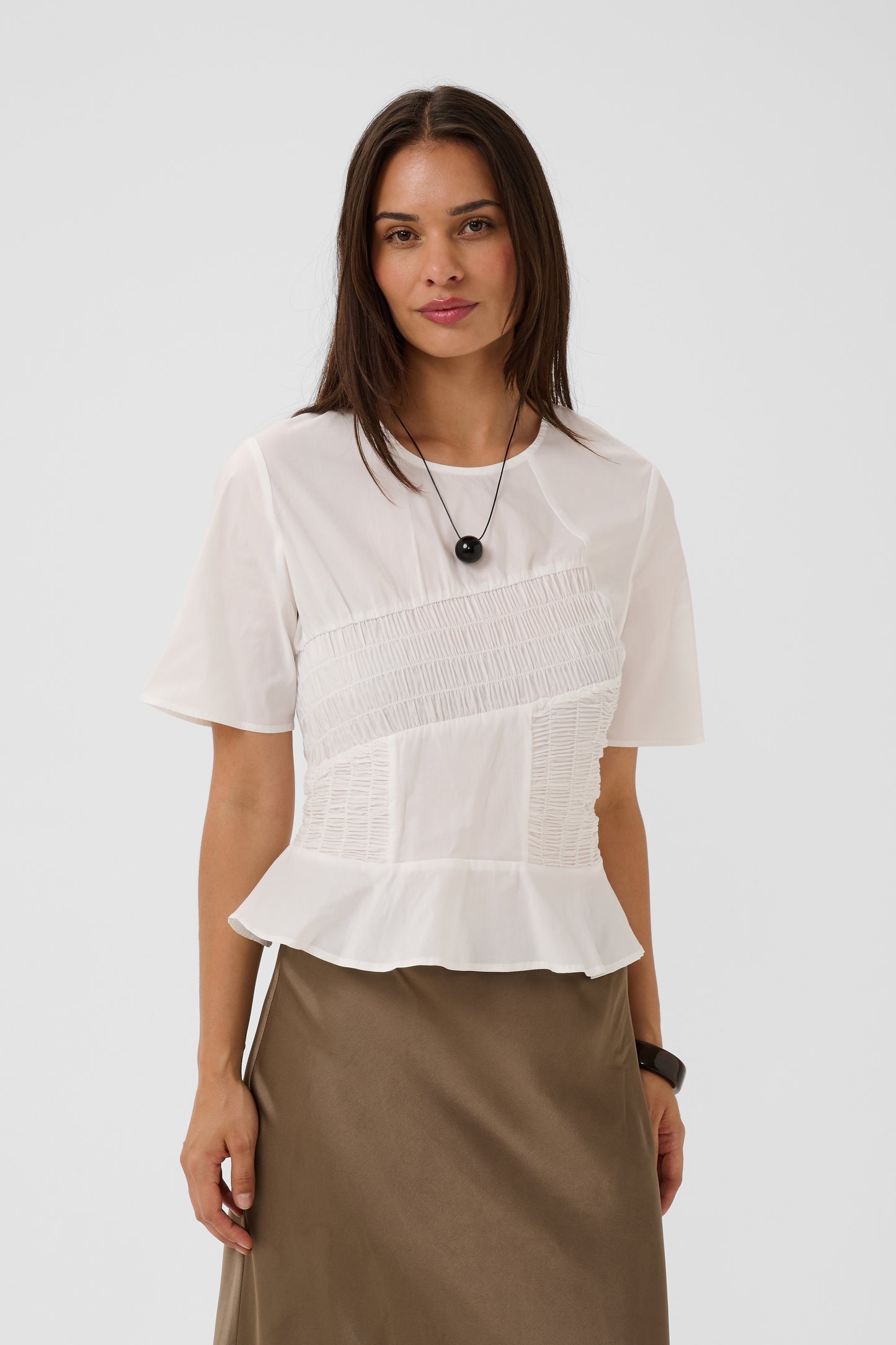 A woman modeling a white short-sleeve blouse with a smocked bodice and peplum hem, paired with a brown skirt, against a light gray studio background.