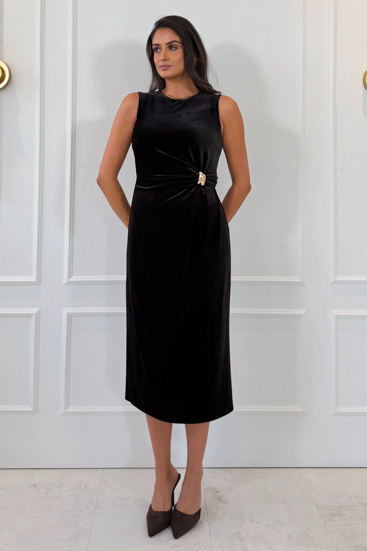 A woman modeling a sleeveless black midi dress with a twisted knot detail at the waist and a small gold accent, standing against a white paneled wall.