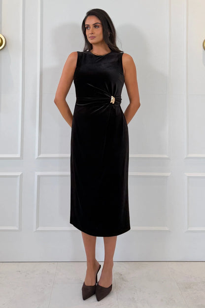 A woman modeling a sleeveless black midi dress with a twisted knot detail at the waist and a small gold accent, standing against a white paneled wall.