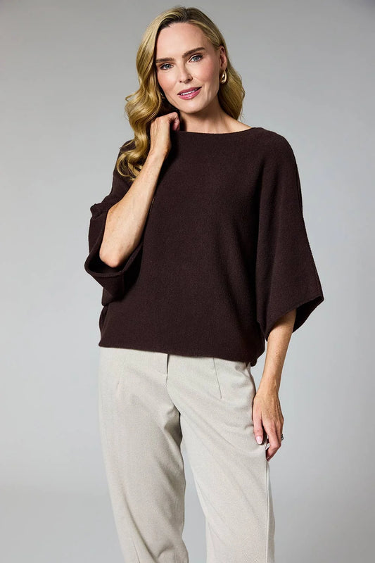 A woman modeling a dark brown boat-neck knit top with wide three-quarter sleeves, paired with light beige trousers, against a solid gray studio background.