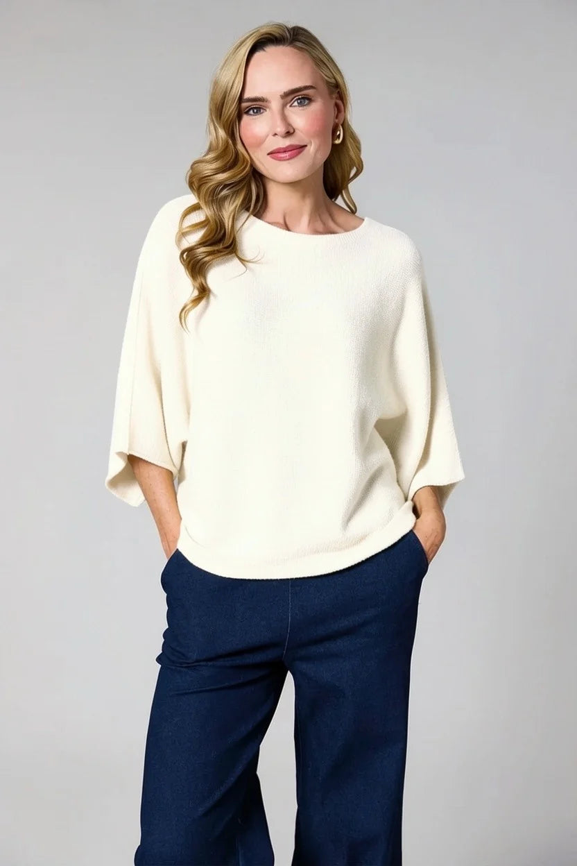 A woman modeling a cream-colored, elbow-sleeve knit top with dark navy wide-leg pants, standing against a neutral light gray studio background.