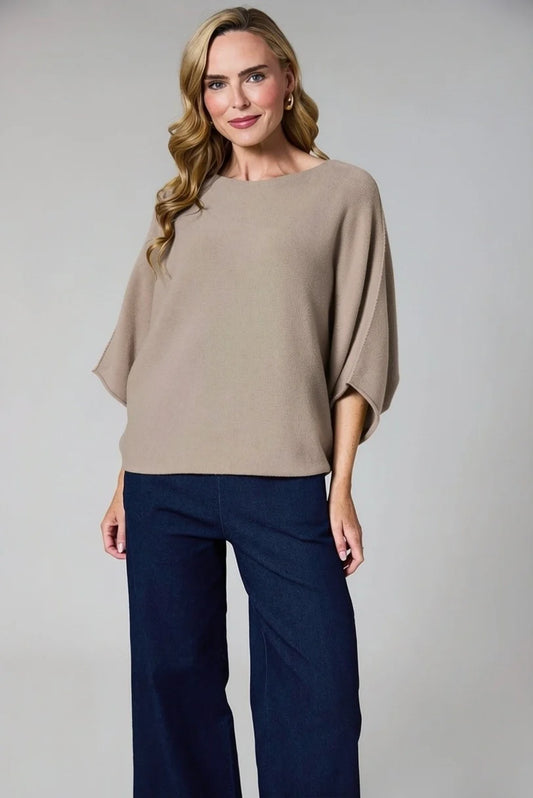 A woman modeling a beige knit top with wide, draped batwing sleeves and dark navy wide-leg pants, photographed against a neutral light gray studio background.