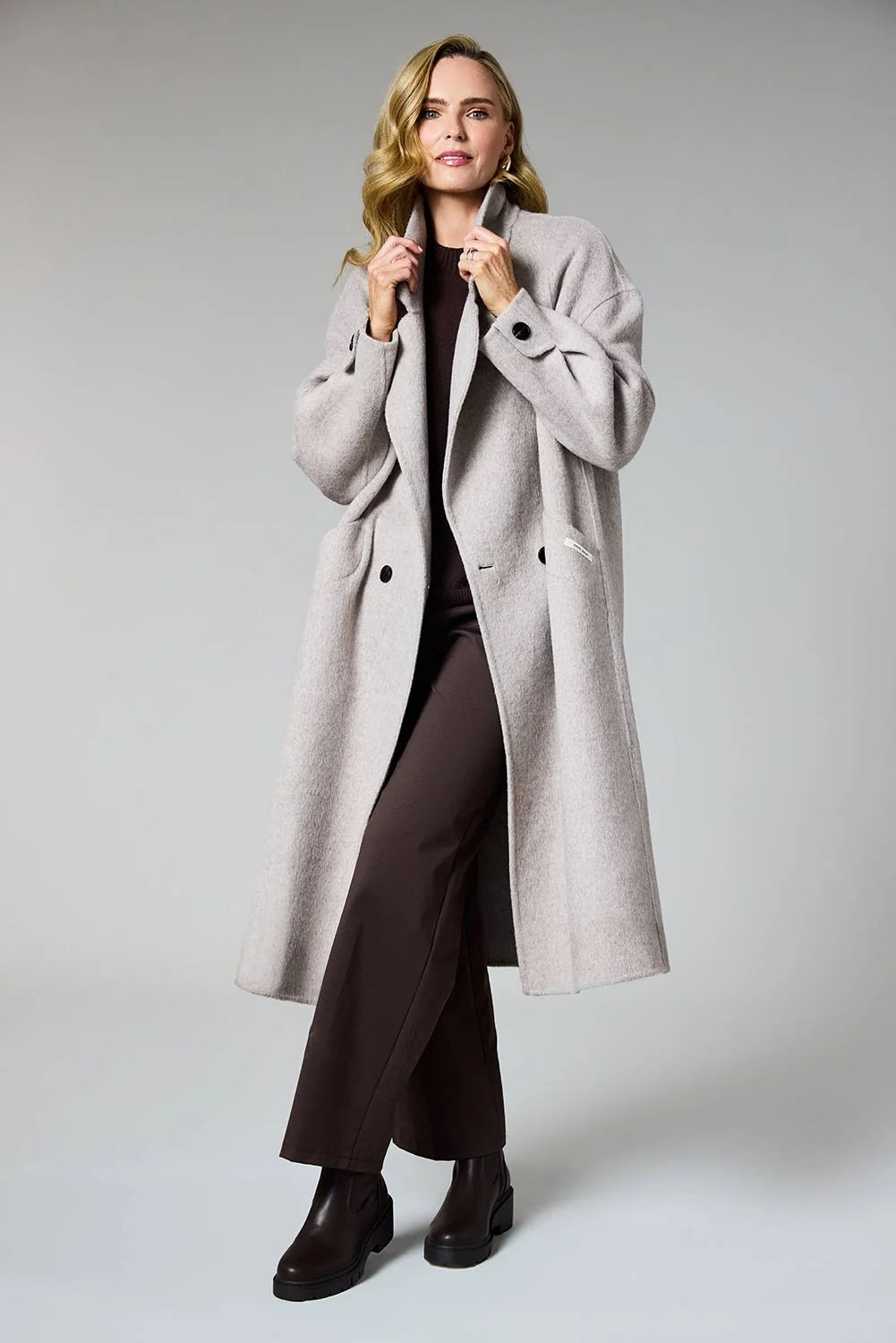A woman modeling a long, light gray double-breasted coat over a dark brown top and wide-leg trousers, standing against a neutral gray studio background.