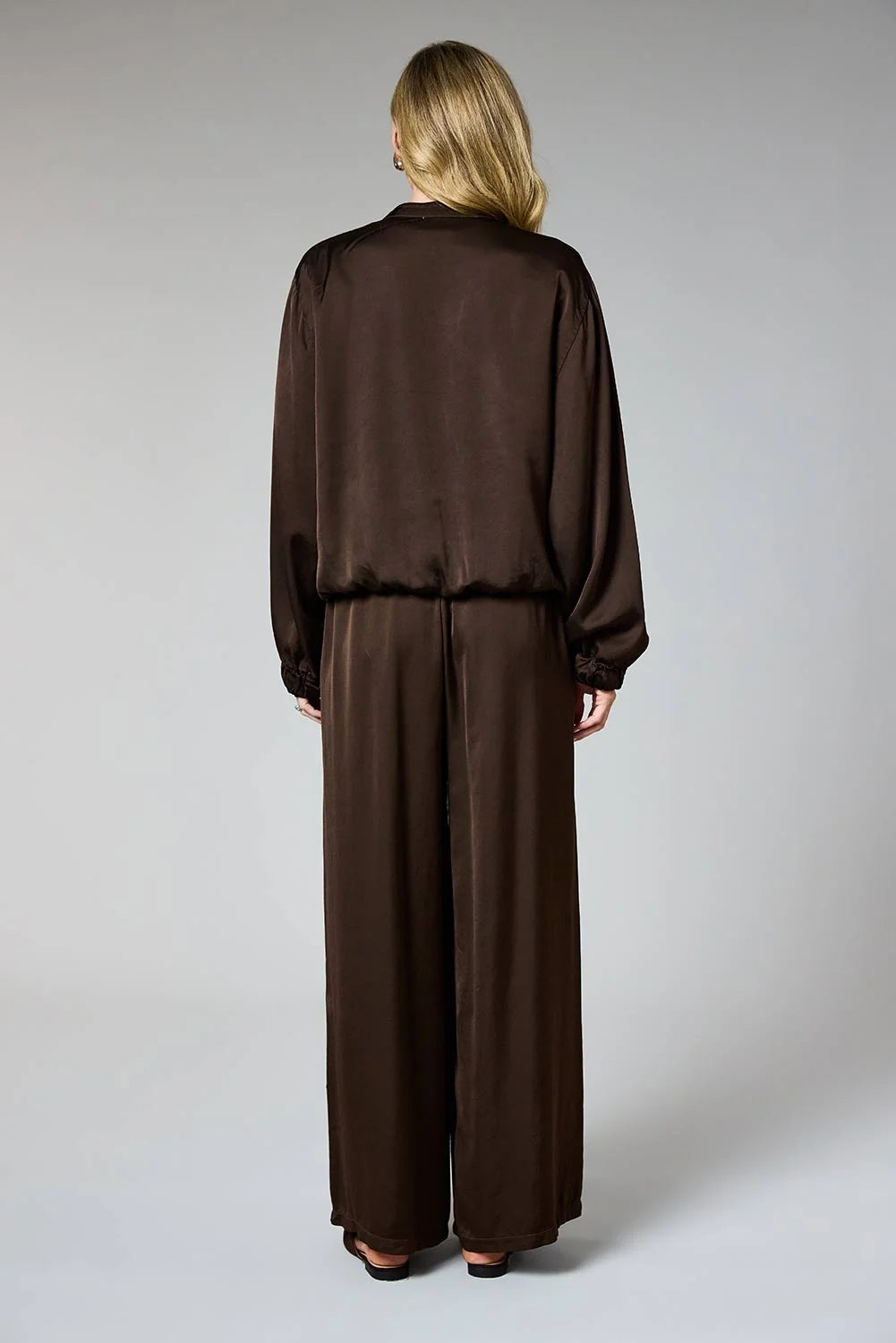 A woman viewed from the back wearing a dark brown satin-like two-piece outfit with a long-sleeve top and wide-leg pants, photographed against a light gray studio background.