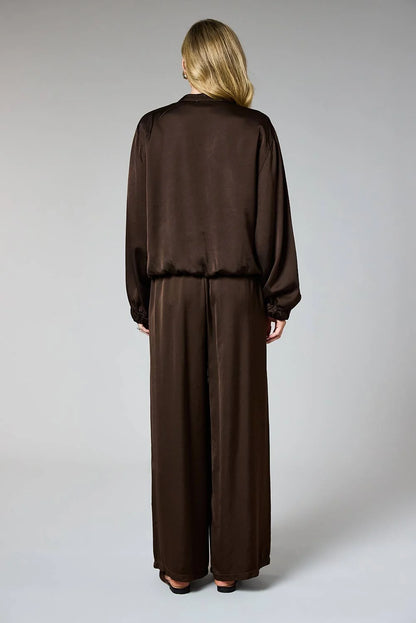 A woman viewed from the back wearing a dark brown satin-like two-piece outfit with a long-sleeve top and wide-leg pants, photographed against a light gray studio background.