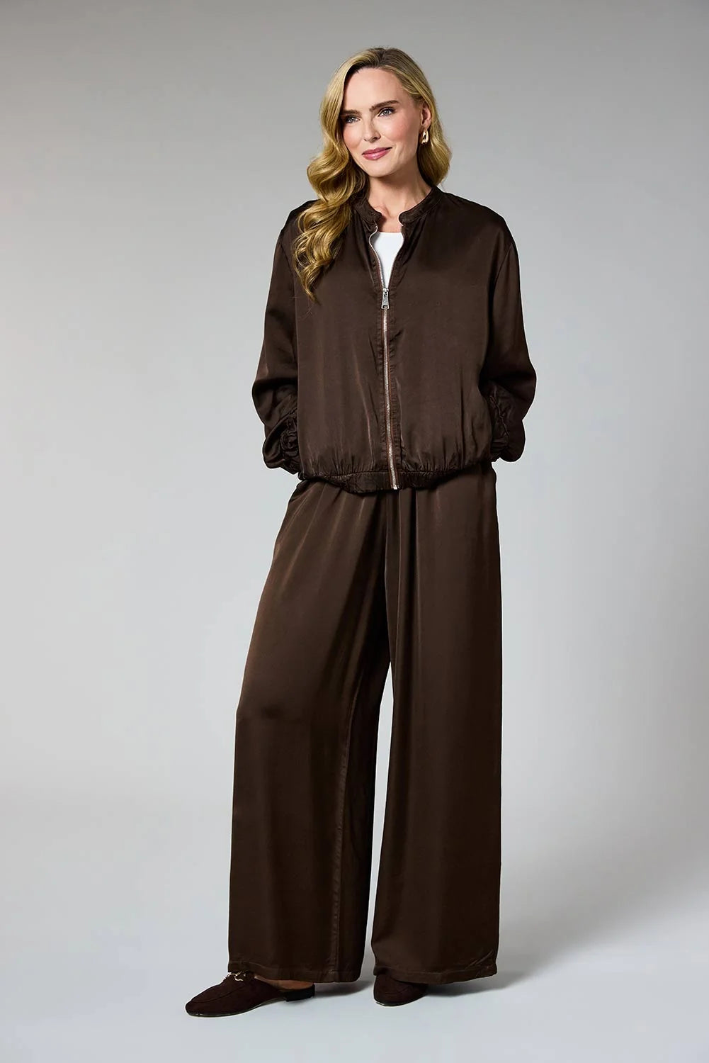 A woman modeling a dark brown two-piece lounge set consisting of a zip-up bomber jacket and matching wide-leg pants, photographed against a neutral gray studio background.