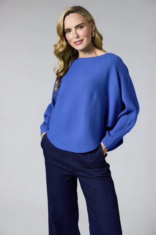 A woman modeling a vibrant blue ribbed knit sweater with balloon sleeves, paired with dark navy wide-leg trousers, against a neutral gray studio background.