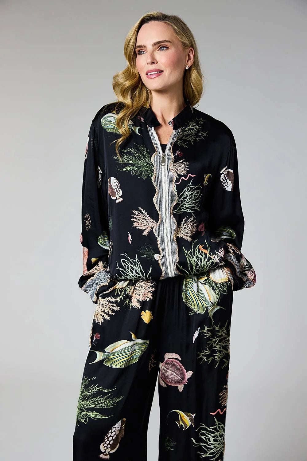 A woman modeling a matching two-piece lounge set with a black satin-like base and a colorful botanical and marine life print, featuring a button-front jacket and wide-leg pants, against a solid gray studio background.