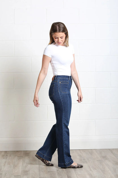 A woman modeling dark blue high-waisted flare jeans with a white t-shirt and leopard print flats, standing against a white brick wall and light wood floor.