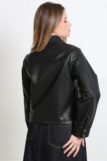 A woman shown from the back wearing a black faux-leather cropped jacket and dark denim skirt against a white brick wall.