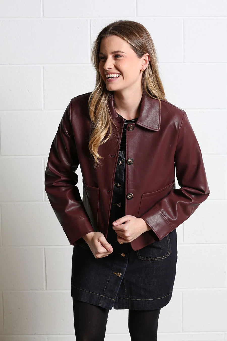 A woman wearing a dark burgundy faux leather jacket over a black top and denim skirt, standing against a white brick wall.