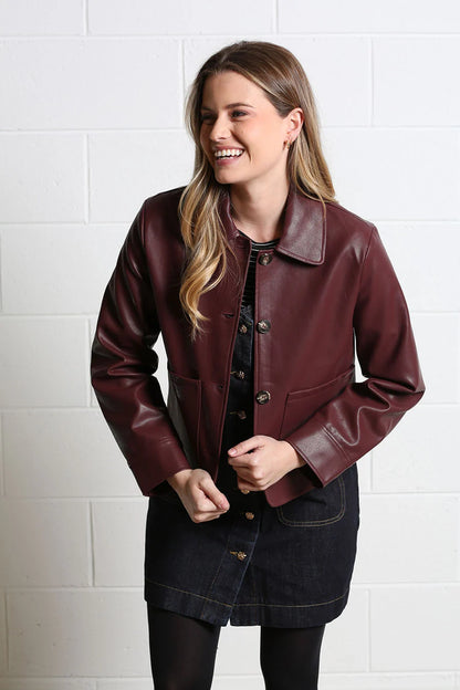 A woman wearing a dark burgundy faux leather jacket over a black top and denim skirt, standing against a white brick wall.