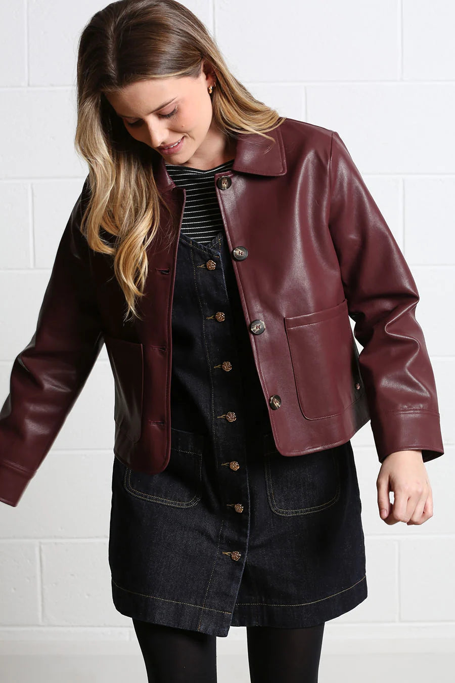 A woman wearing a cropped, short-sleeved burgundy faux-leather jacket over a dark denim button-front dress and black tights, standing against a white brick wall.