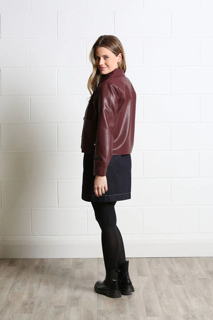 A woman stands in profile wearing a cropped burgundy leather jacket, a dark navy mini skirt, black tights, and black ankle boots, set against a white brick wall and light wood floor.