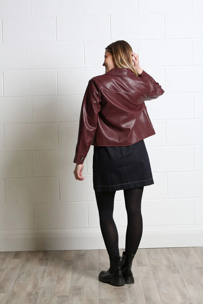 A woman stands with her back to the camera wearing a cropped, shiny burgundy faux-leather jacket, a dark denim skirt, black tights, and black ankle boots, set against a white painted brick wall and light wood floor.