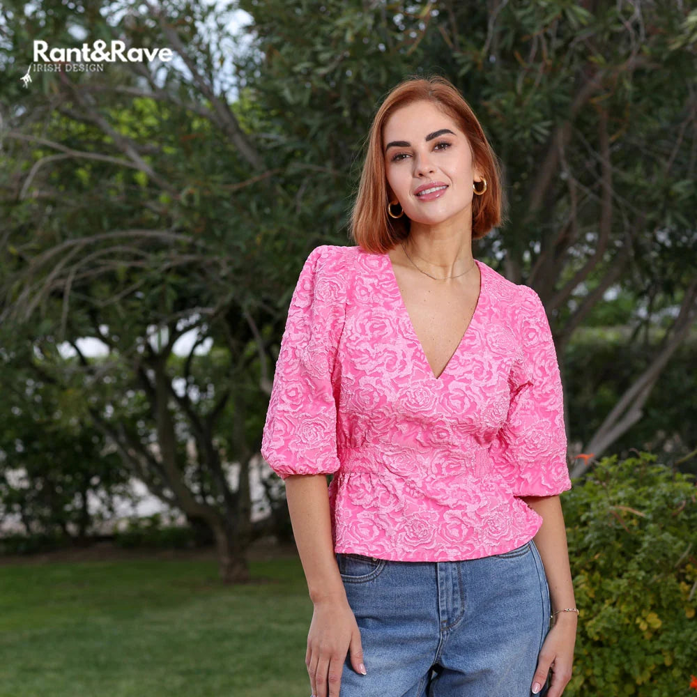 Woman wearing a pink patterned top and blue jeans standing outdoors with greenery in the background
