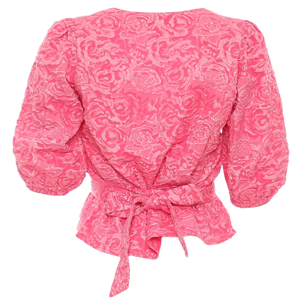 A pink long-sleeve blouse with a textured rose pattern, featuring a wrap-style back with a tie belt and puffed sleeves, shown against a plain white background.