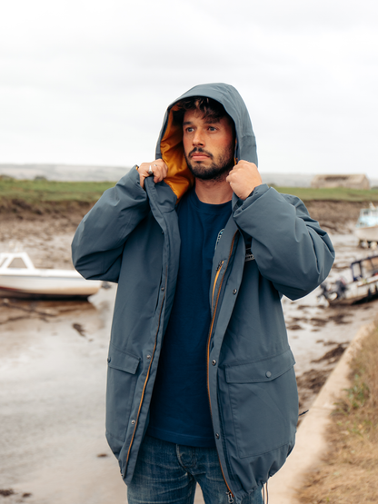 A man wearing a slate blue hooded raincoat with a bright orange interior lining, standing on a muddy shoreline with boats in the background.