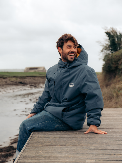 A man wearing a dark slate blue hooded rain jacket and blue jeans sits on a wooden boardwalk outdoors on an overcast day.