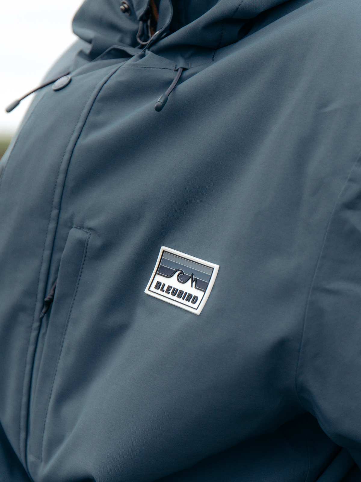 Close-up of a person wearing a slate blue hooded jacket, featuring a white and blue rectangular 'BLEURIND' logo patch on the chest.