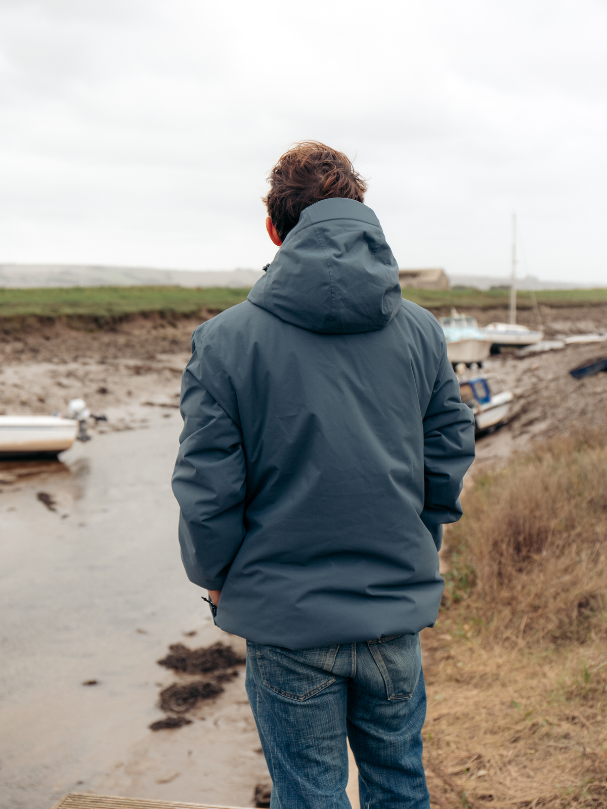 A man viewed from behind wearing a slate blue hooded jacket and blue jeans, standing on a muddy shoreline next to a small body of water with boats in the background.