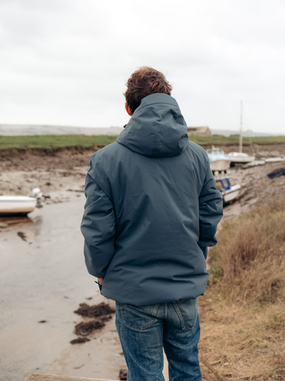 A man viewed from behind wearing a slate blue hooded jacket and blue jeans, standing on a muddy shoreline next to a small body of water with boats in the background.