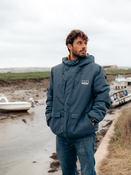 A man wearing a navy blue Patagonia hooded jacket and blue jeans stands on a muddy shoreline with boats and an overcast sky in the background.