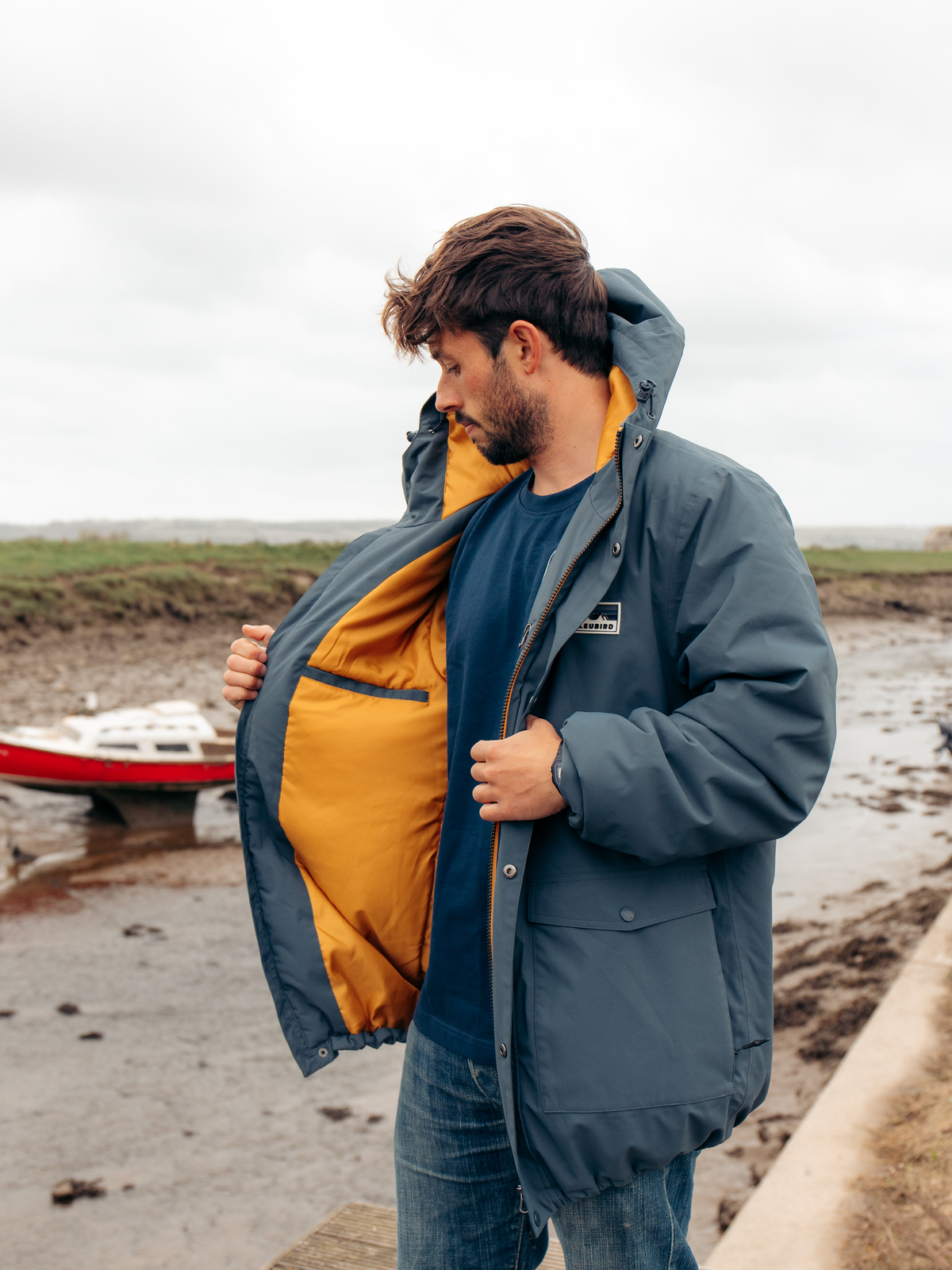 A man wearing a slate blue hooded puffer jacket with a bright yellow-orange interior lining, standing outdoors near a muddy shoreline with a small boat in the background.