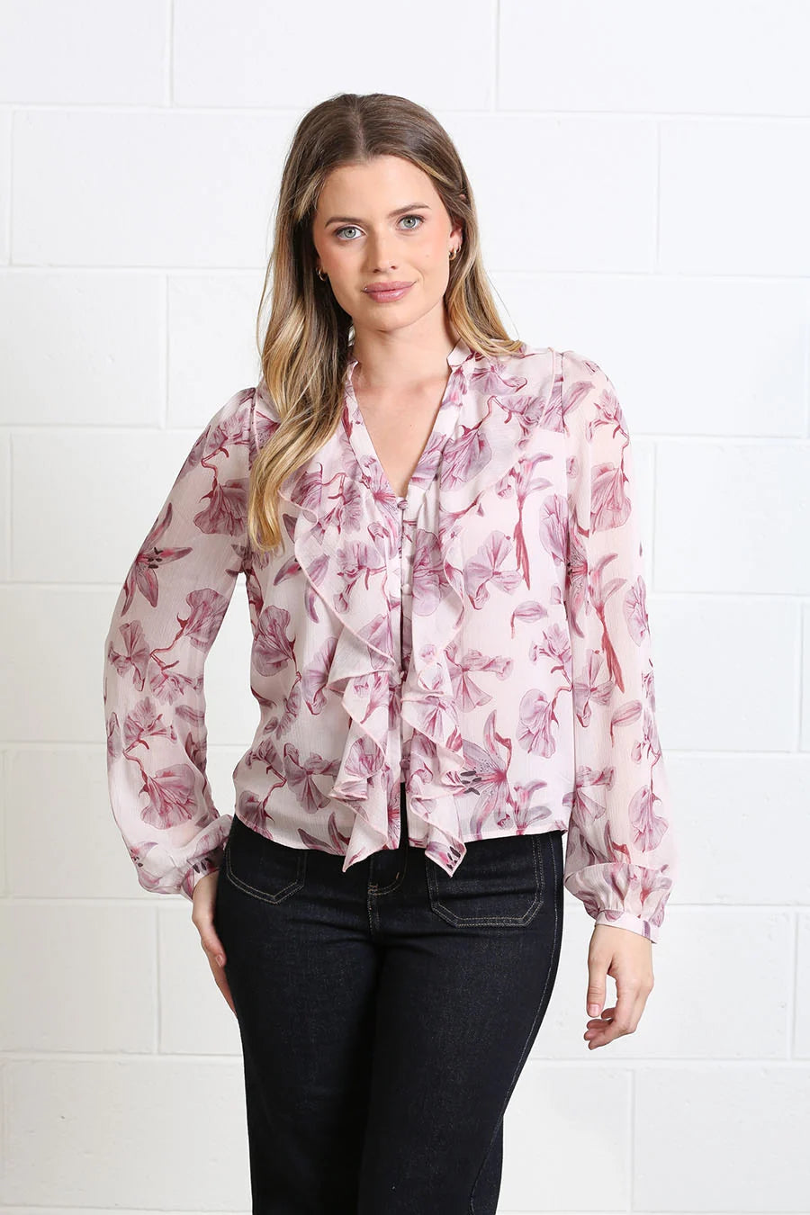 A woman modeling a sheer long-sleeve blouse with a pink and mauve floral print, ruffled trim, and a tie-neck detail, paired with dark jeans against a white brick wall.