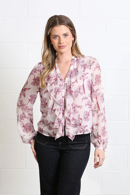 A woman modeling a sheer long-sleeve blouse with a pink and mauve floral print, ruffled trim, and a tie-neck detail, paired with dark jeans against a white brick wall.