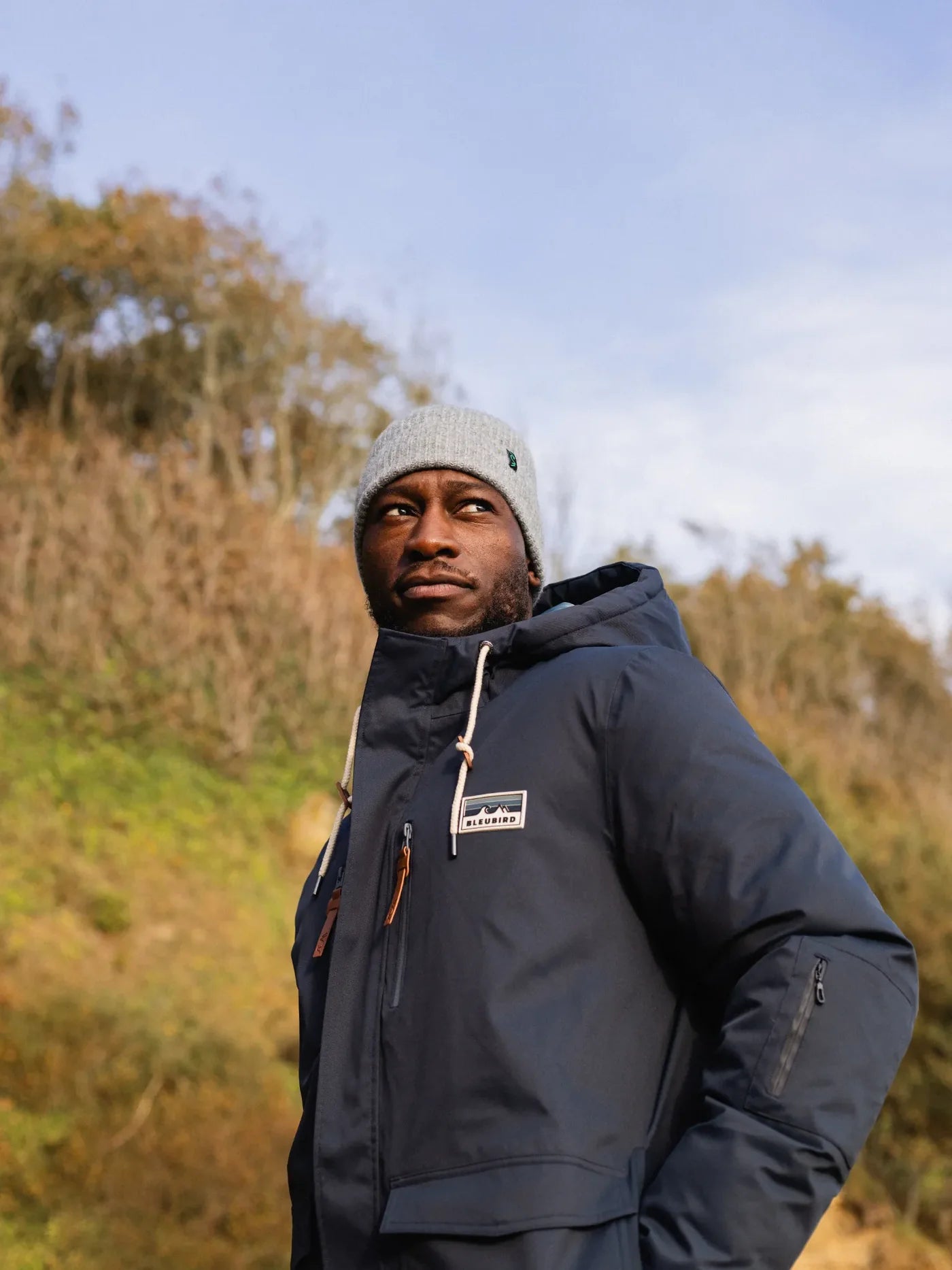 Man wearing a blue jacket and green pants standing on a rocky beach.