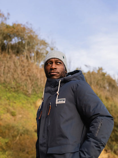 Man wearing a blue jacket and green pants standing on a rocky beach.