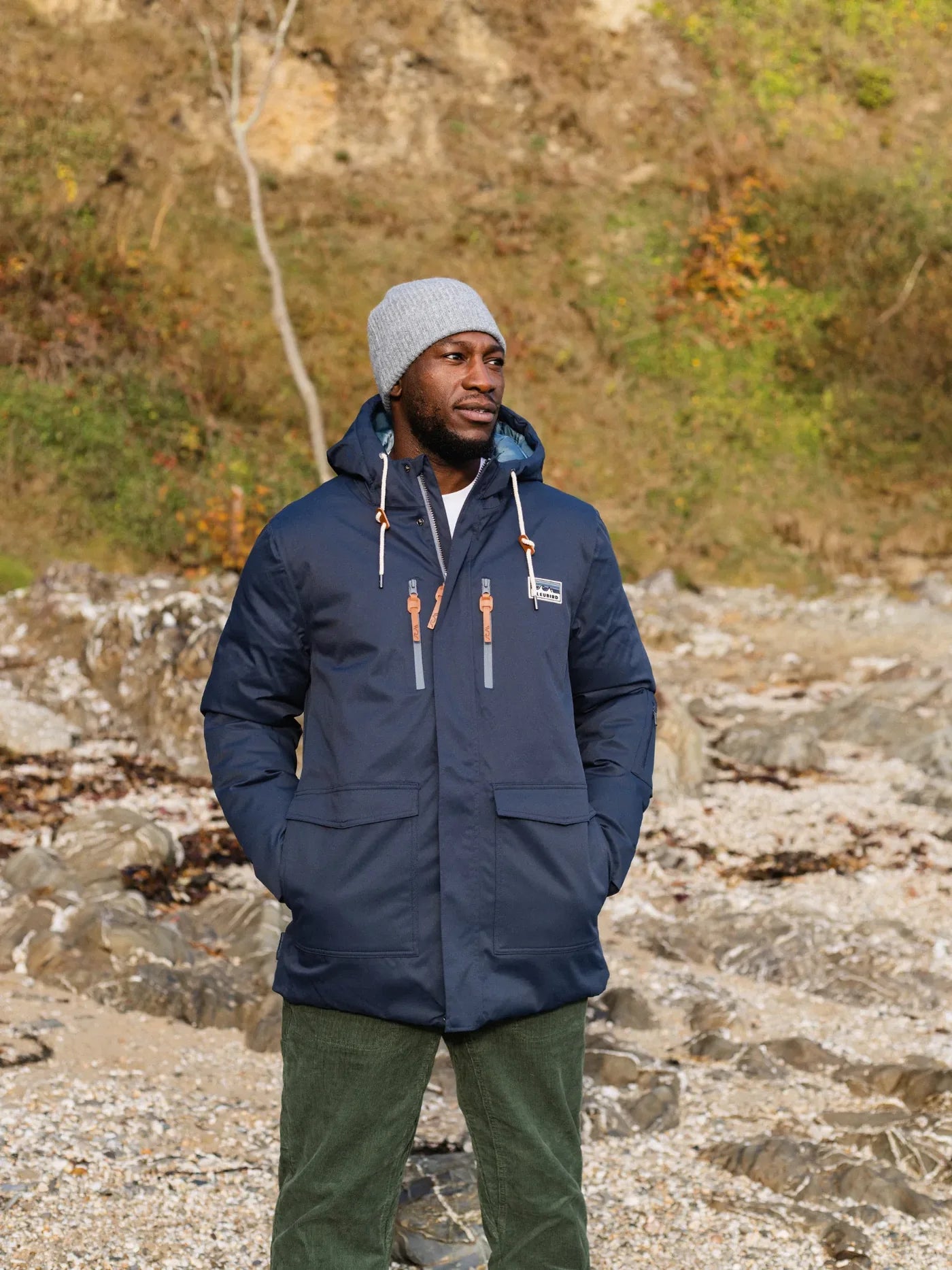 Man wearing a blue jacket and gray beanie standing on a rocky terrain with a natural background.