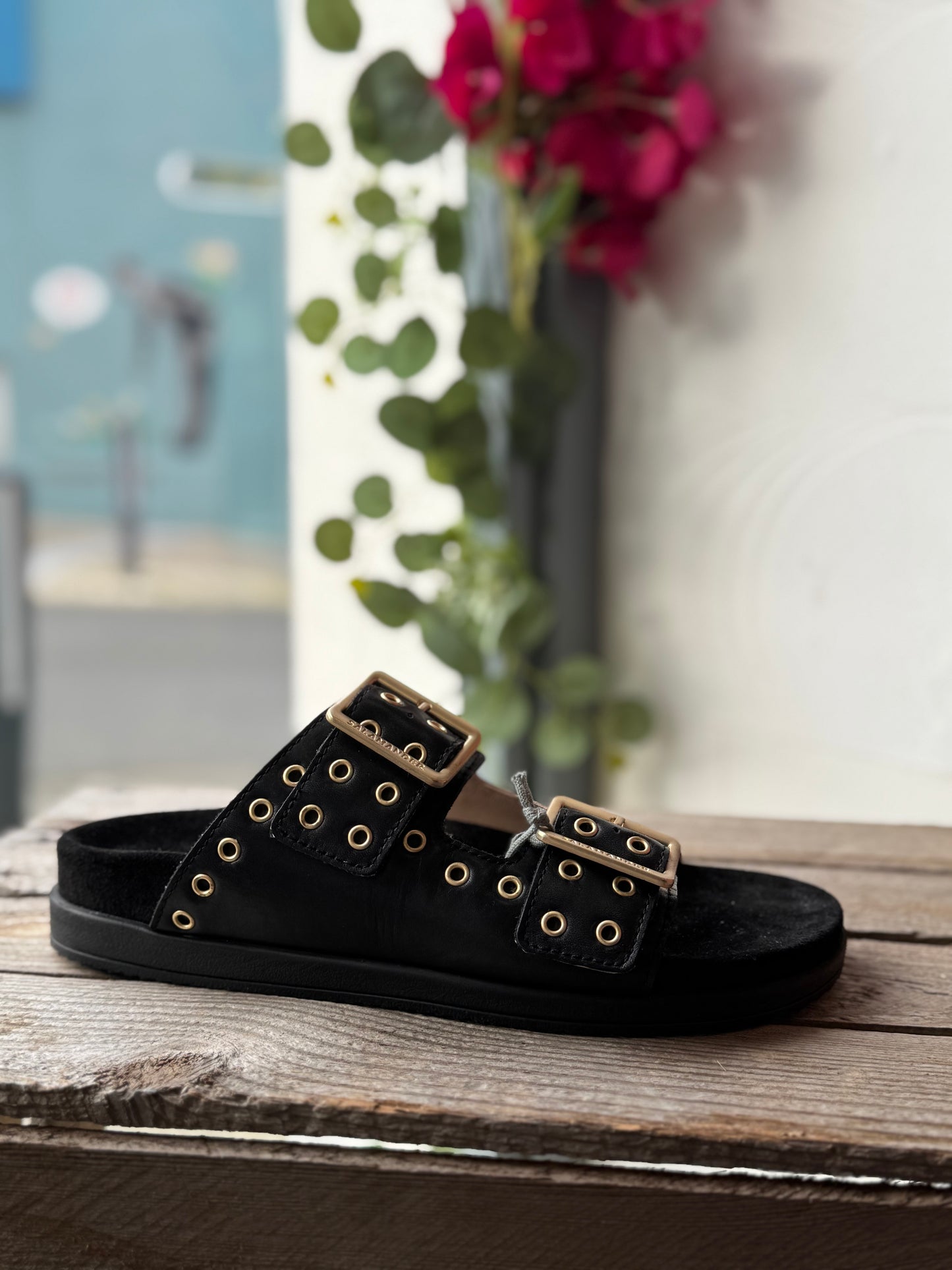 Black sandal with gold buckles on a wooden surface with flowers in the background