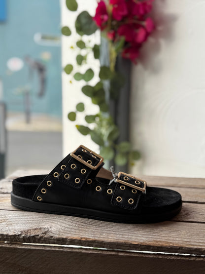 Black sandal with gold buckles on a wooden surface with flowers in the background