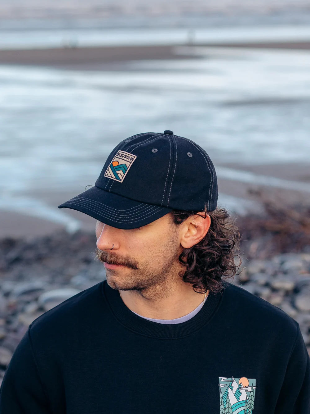 Man wearing a black cap and sweater with a logo, standing by a body of water.