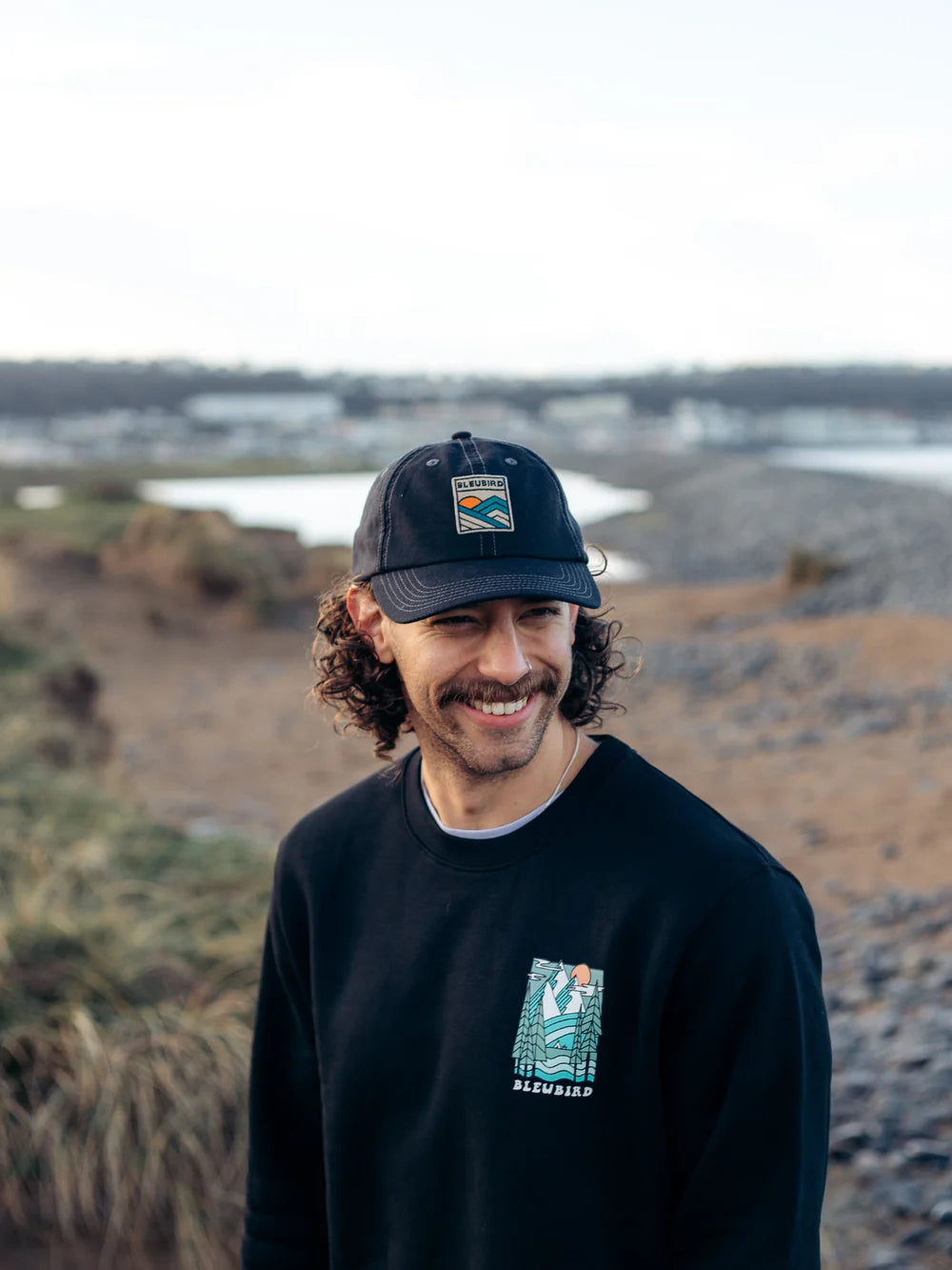 Man wearing a black cap and sweatshirt with logos, standing in a natural landscape.