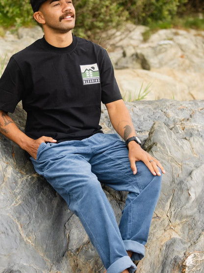 Man sitting on rocks wearing a black t-shirt with a logo and blue jeans.