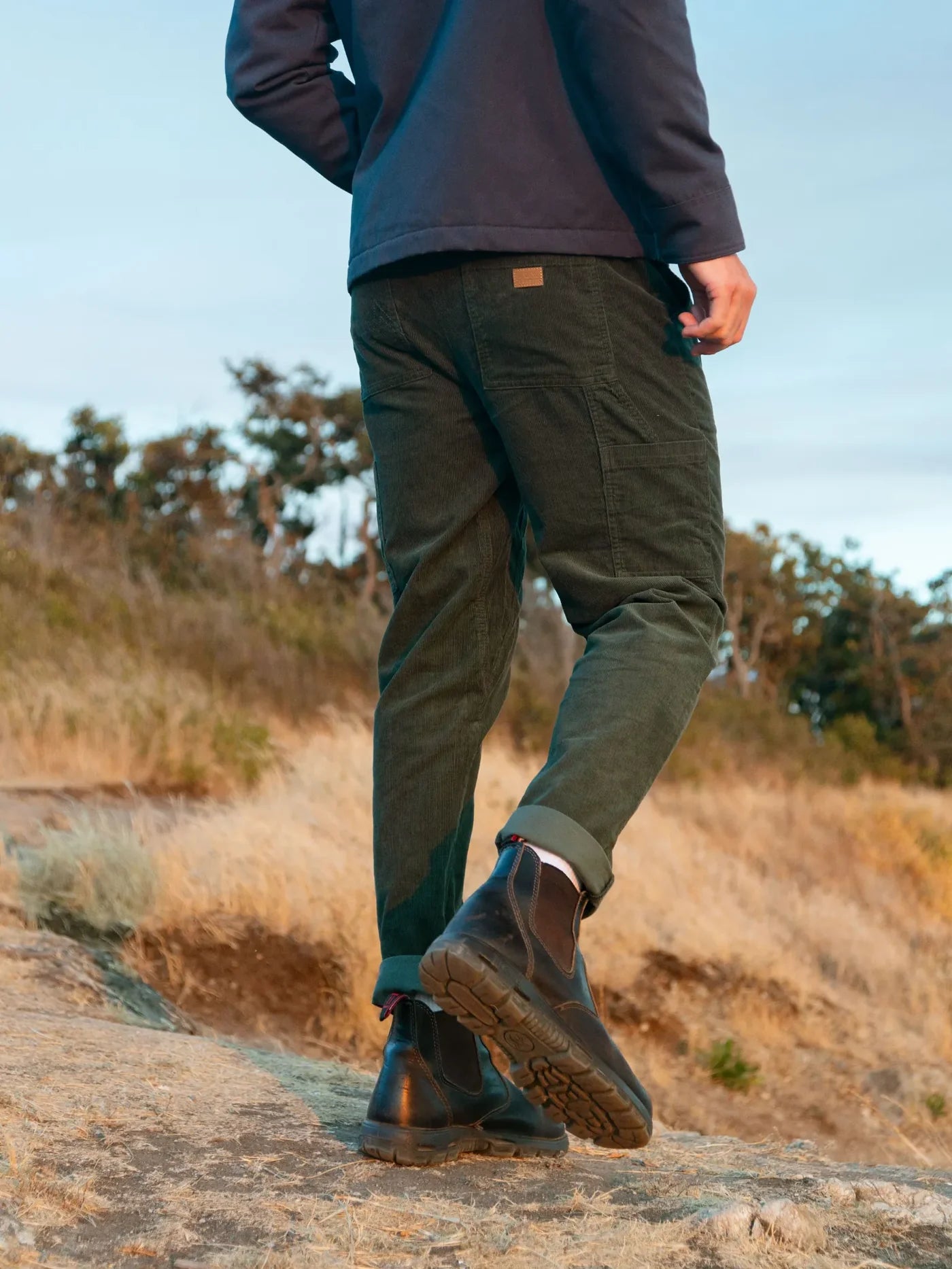 Person wearing dark jacket, green pants, and brown boots walking on a rocky path with trees in the background.