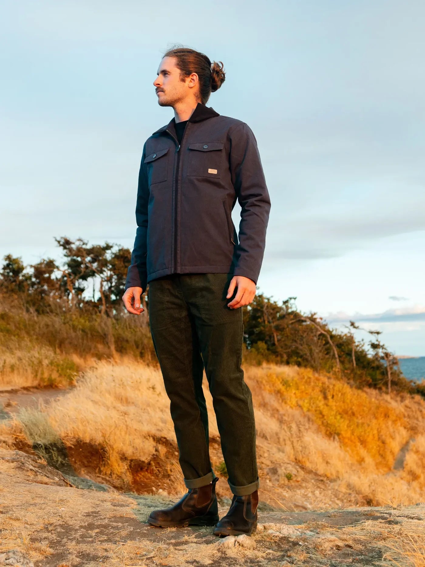 Man standing on a trail wearing a dark jacket and green pants, with a scenic background.