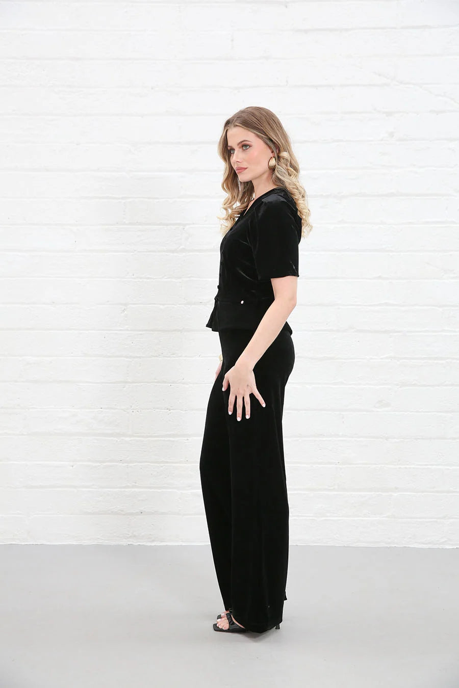 A woman modeling a black velvet short-sleeve top and matching wide-leg trousers, standing in profile against a white brick wall.