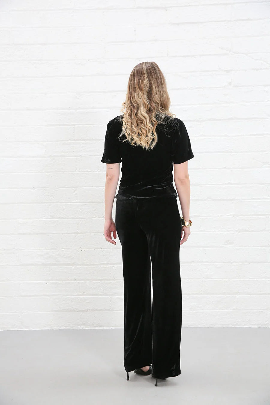 A woman with blonde hair is shown from behind wearing a black velvet short-sleeve top and matching wide-leg trousers, standing against a white brick wall.