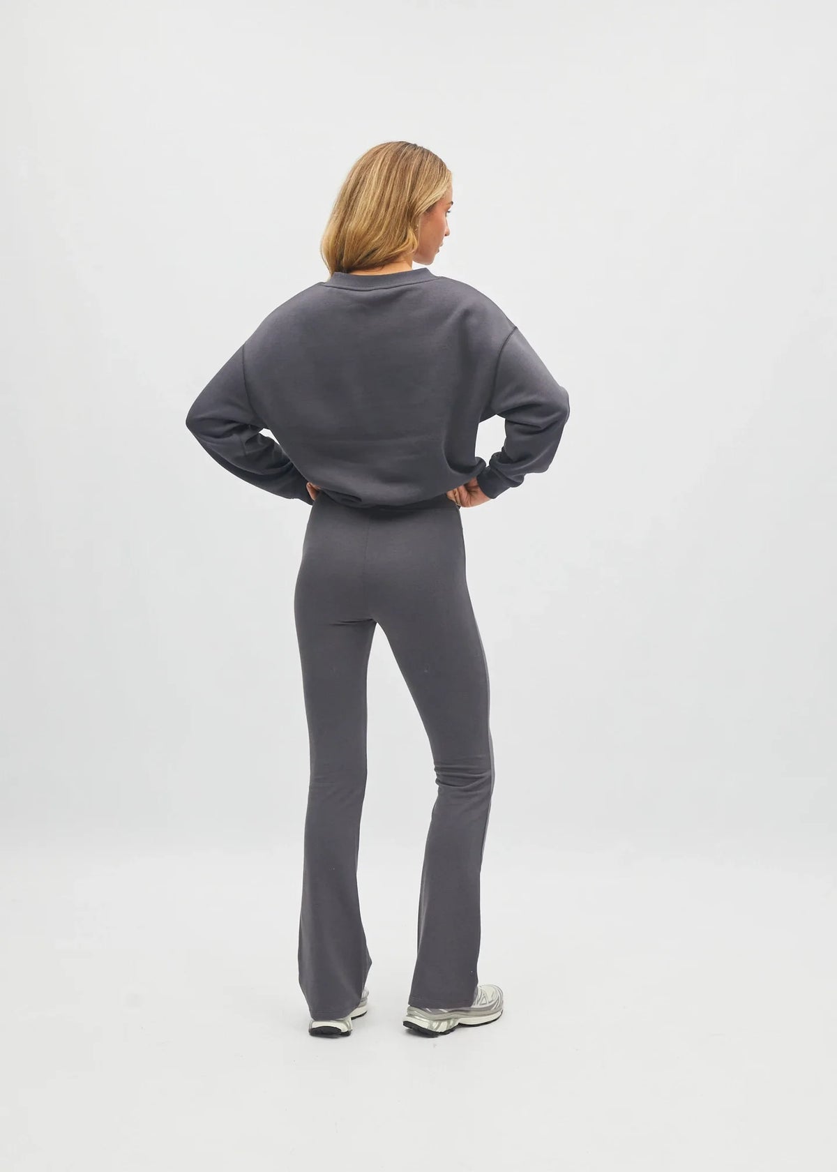 A woman stands with her back to the camera wearing a dark gray sweatshirt and matching flared leggings, paired with gray sneakers, against a light gray studio background.
