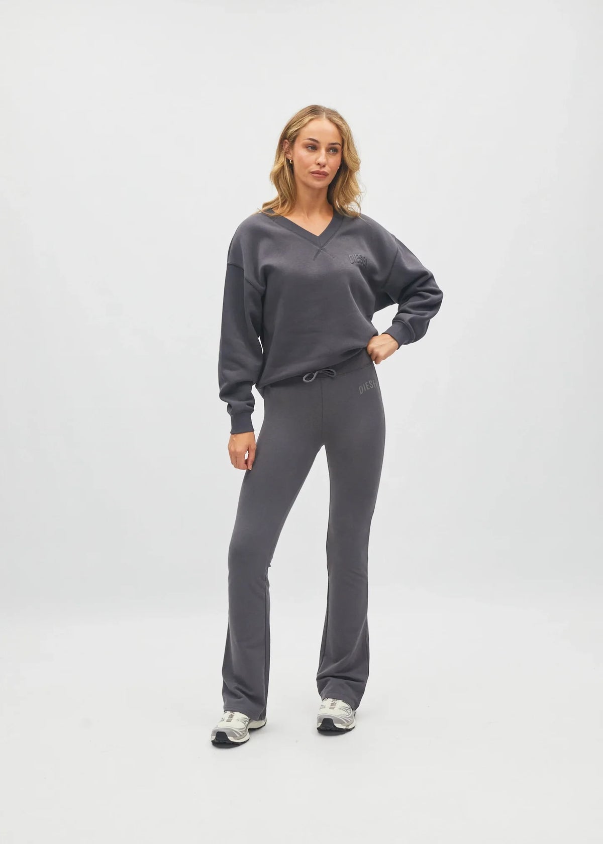 A woman modeling a dark gray two-piece loungewear set consisting of a V-neck sweatshirt and matching drawstring pants, standing against a light gray studio background.
