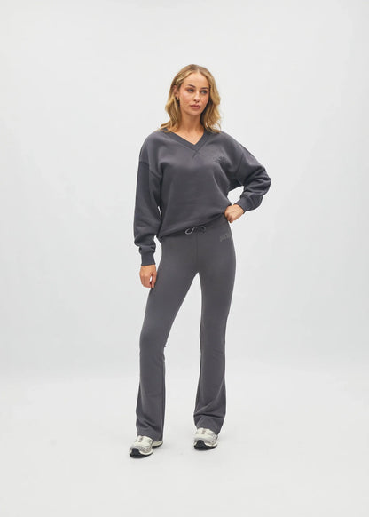 A woman modeling a dark gray two-piece loungewear set consisting of a V-neck sweatshirt and matching drawstring pants, standing against a light gray studio background.